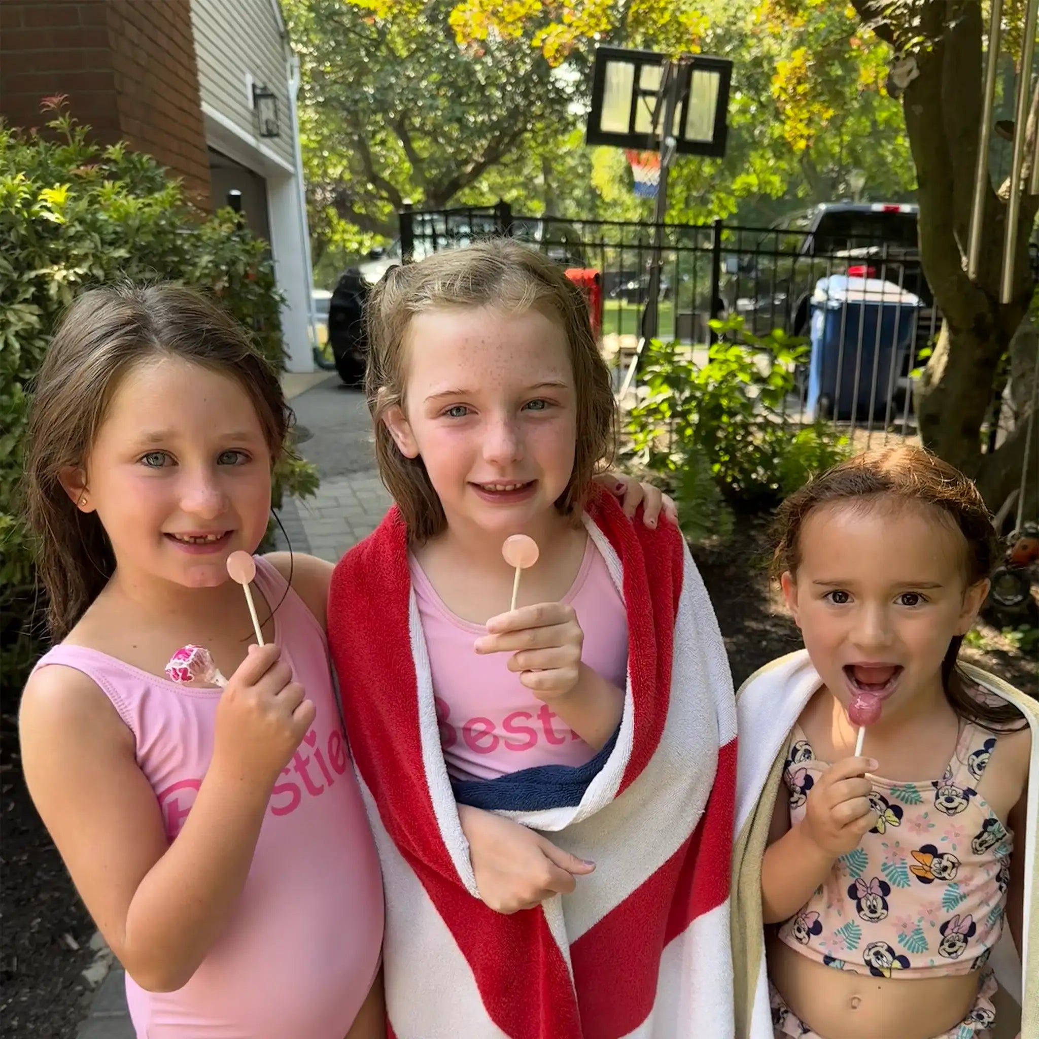 Three young girls outdoors, wrapped in towels, holding lollipops.