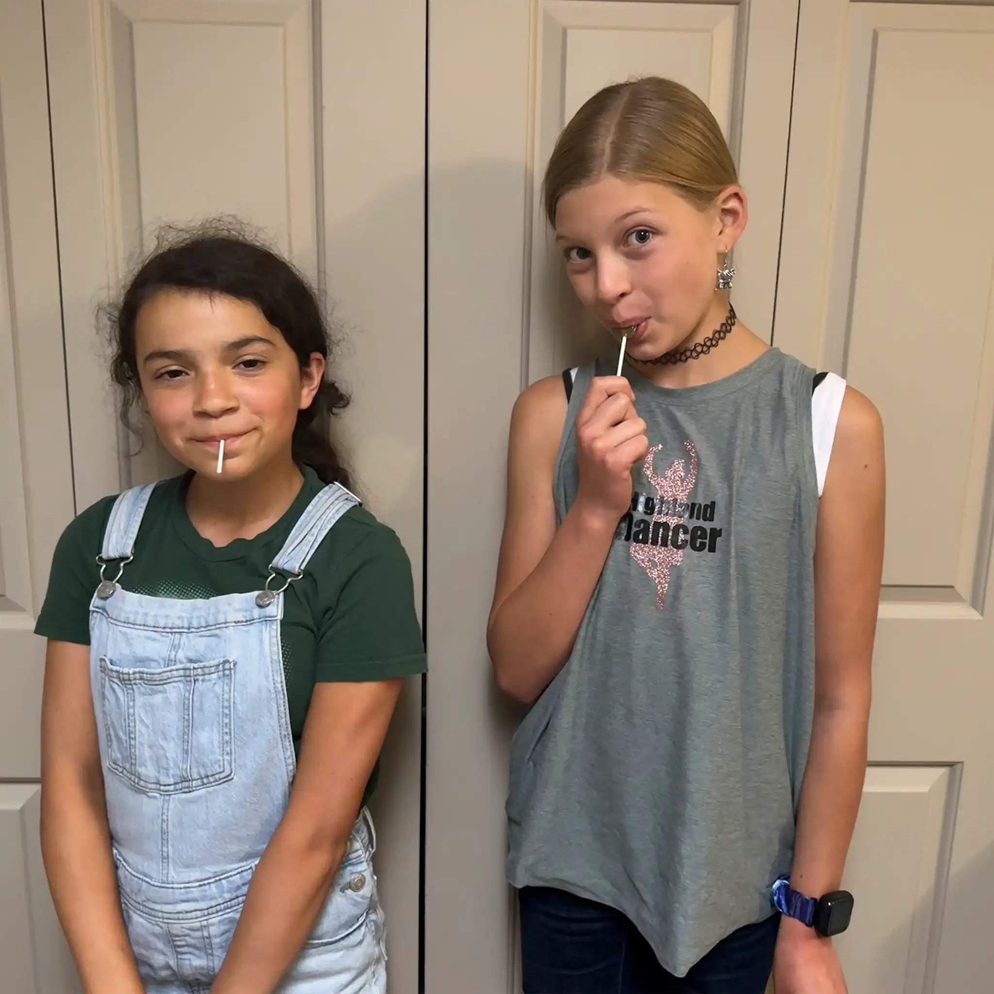 Two young girls standing in front of a door, one wearing overalls and a green shirt, the other in a gray sleeveless top.