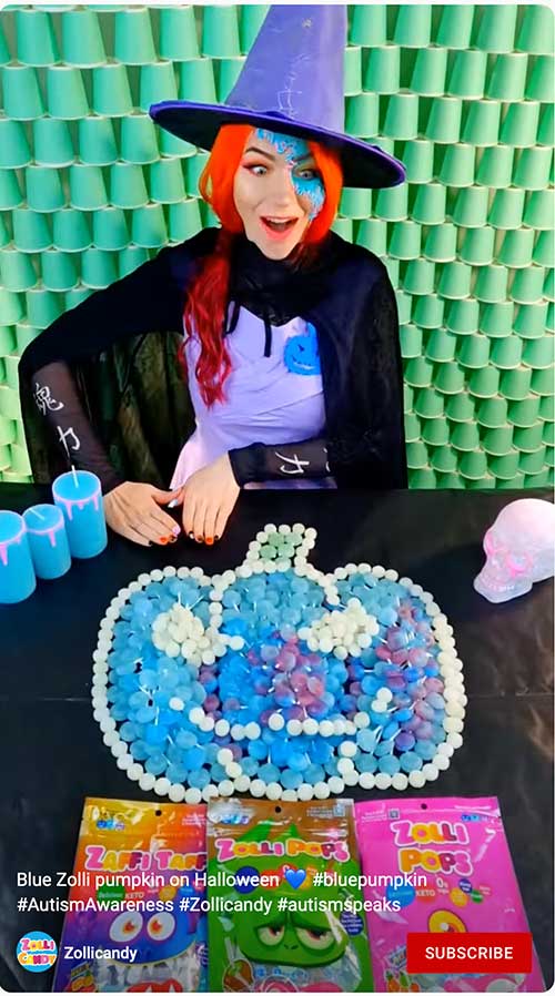 A woman in a witch costume with a blue pumpkin made of Zolli Pops on a table, promoting Autism Awareness for Halloween.