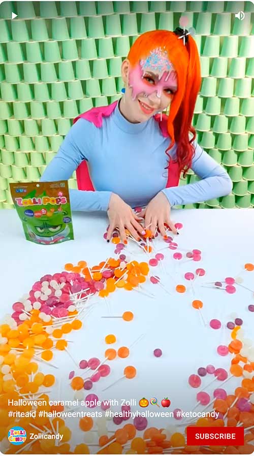 A woman with vibrant makeup and orange hair arranges colorful Zolli Pops on table in a Halloween-themed video thumbnail