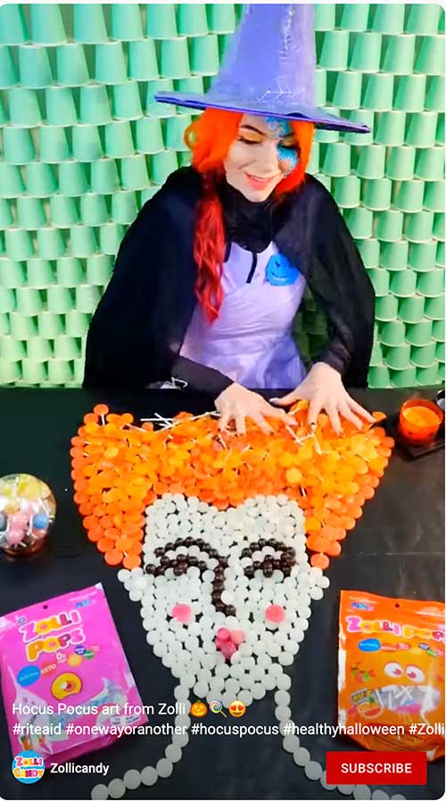 Girl dressed as a witch creating a candy art portrait with orange hair surround by candy bags and a colorful backdrop