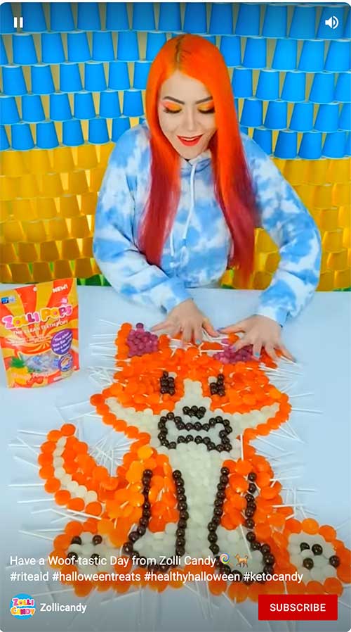 Person with orange hair arranging candy into a fox shape on a table, with a colorful backdrop and candy box nearby.